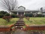 Single-story brick house with a covered front porch, two American flags, and steps leading up through a grassy yard with bushes. The sky is overcast and leafless trees are visible in the background.
