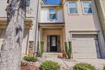 A two-story townhouse with beige siding, a black front door, one-car garage, small shrubs, and rocks lining the walkway, photographed on a sunny day.