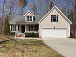 A beige two-story house with white trim, black shutters, a covered front porch, and an attached two-car garage. The house is surrounded by leafless trees and has a driveway and front lawn.