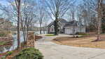 A two-story house with a three-car garage sits at the end of a curved driveway beside a lake. Leafless trees surround the property, and a stone mailbox stands near the driveway entrance.