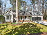 A single-story house with light-colored siding, a front porch, black double garage doors, and a sloped roof sits among tall trees with a lawn, driveway, and garden beds in front.