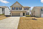 Two-story beige suburban house with black shutters, a double garage, and a concrete driveway. The front yard has dry grass, a small tree, and minimal landscaping. Neighboring houses are visible on both sides.