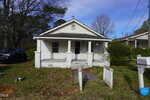 A small, single-story white house with a covered front porch and chipped paint, set back from the street with an overgrown front yard. A black truck is parked to the left, and a blue "For Sale" sign is on the right.