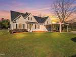 Single-story house with beige siding, black roof, covered front porch, and attached carport, set on a large green lawn at sunset with a bare tree to the right and shrubs along the front of the house.