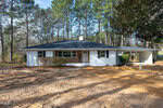 A single-story white house with a dark roof, front porch, and attached carport sits in a clearing surrounded by tall trees and patchy dirt yard. Sparse bushes line the front of the house.