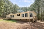 A single-story, light yellow manufactured home with black shutters and a brick foundation, surrounded by tall pine trees and set on a dirt and grassy clearing. The house has two small wooden porches with steps.