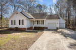 Single-story white house with a front porch, attached garage, gable roof, brick foundation, and concrete driveway. The yard is mostly bare with some grass, surrounded by trees under a partly cloudy sky.