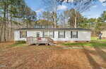 A single-story white manufactured home with black shutters and a small wooden deck, set in a grassy yard surrounded by tall trees under a partly cloudy sky.