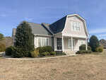 A beige two-story house with a gambrel roof, white trim, front porch, and shrubbery in the yard, set against a blue sky with some neighboring houses visible in the background.