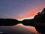 A serene lake at dusk reflects a colorful sky with pink, orange, and purple clouds. Silhouetted trees frame the horizon, and a small crescent moon is visible in the upper left corner.