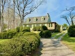 A two-story house with dormer windows and a steep roof sits on a slightly elevated lawn. Trimmed green bushes line the driveway, and tall bare trees surround the property under a clear sky.