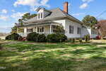 A white, single-story house with a gray roof, front porch, brick steps, and multiple windows is surrounded by green grass, shrubs, and trees under a partly cloudy sky.