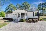 A single-story white manufactured home with black shutters, a front porch with steps, and neatly trimmed bushes lines the front. Gravel driveway and green trees are visible in the background under a blue sky.