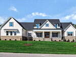 A large, two-story suburban house with light siding, stone accents, a dark shingle roof, multiple gables, and a wide front porch with columns. The front yard is grassy with minimal landscaping.