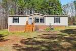 A single-story manufactured home with beige siding and a brick foundation sits on a grassy lot. There are wooden steps leading to the front door and tall trees in the background.