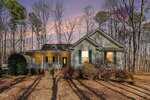 A single-story house with a large front porch, green shutters, and a glass front door is surrounded by leafless trees at sunset. Warm lights illuminate the porch and interior, with fallen leaves covering the yard.