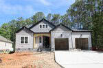 A newly built single-story house with gray siding, white trim, a covered front porch, and a double garage. The driveway and yard are unfinished, and the house is surrounded by tall trees.