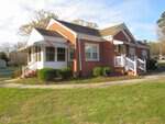 A one-story red brick house with two white stairways, striped awnings over the entrances and windows, and a screened porch, set on a curved walkway with trimmed bushes and grass in the foreground.