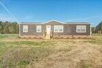 A single-story, gray manufactured home with white trim and a small front porch sits on a grassy, partially bare yard under a clear blue sky. There are trees in the background.