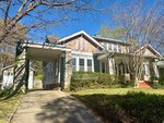 A two-story house with a brick and light blue exterior, white trim, and a covered carport on the left. There are shrubs and trees in the front yard and a driveway leading to the carport.