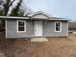 A small single-story house with gray vinyl siding, a white front door, two windows, and a small covered entryway. The yard is mostly bare dirt with patches of grass. Trees are visible in the background.