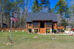 A small log cabin with a front porch sits in a grassy yard with shrubs and a U.S. flag on a pole. The background features tall trees and a clear blue sky, with a shed and parked vehicle visible to the right.