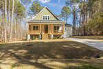 A two-story house with green and beige siding, a covered front porch with wooden railings, and steps leading to the entrance, situated on a grassy lot surrounded by tall trees and a concrete driveway.