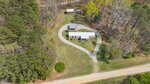 Aerial view of a single-story house with a gray roof, a circular gravel driveway, a small outbuilding, and a larger shed, surrounded by trees and grassy areas near a dirt road.