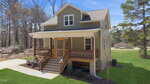 A two-story house with beige siding, a covered front porch with wooden railings, and a set of stairs. The house is surrounded by grass, trees, and a paved walkway, with a detached garage in the background.