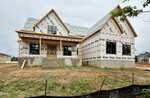 A two-story house under construction, covered in weatherproof barrier wrap with exposed wooden framing, sits on a concrete foundation with unfinished steps and a dirt yard in front.