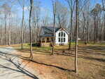 A small white house with large windows and a front porch sits among tall leafless trees, surrounded by a dirt and grass yard, with a gravel driveway in the foreground.