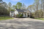 Single-story white house with black roof and a covered front porch, surrounded by trees and greenery; located next to a fenced yard and paved driveway on a clear day.
