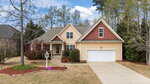 A beige and red suburban house with a two-car garage, a driveway, small front porch, landscaped yard with mulch, shrubs, and trees, under a partly cloudy sky.