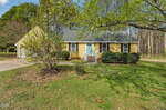 A yellow single-story house with white trim, a teal front door, and a front porch. A detached garage is on the left, and the yard has grass, shrubs, and large trees with green leaves.