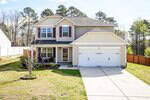 Charming two-story suburban home at 820 Pitch Pine Drive, Creedmoor, with tan siding, red shutters, a double garage, and a cozy porch with two chairs. The house is surrounded by grass, trees, and a wooden fence.