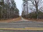 A gravel driveway extends from a paved road into a wooded area with tall trees on both sides. The trees have sparse leaves, suggesting late autumn or winter. The sky is overcast.