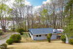 A single-story house with a gray roof sits on a grassy lot surrounded by trees. A gravel driveway leads to the house, and a lake is visible through the trees in the background under a partly cloudy sky.