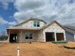 A partially constructed house with visible framing, house wrap, and unfinished roof. The front porch and garage are not yet completed, and the ground is bare dirt. The sky is mostly cloudy.