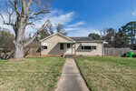 Single-story beige house with a dark roof, white trim, and two awnings over the front windows. A concrete walkway leads to the front door. The yard has grass, a large leafless tree, and is enclosed by a wooden fence.