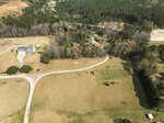 Aerial view of a rural area with a winding dirt road, a large house with a driveway on the left, several trees, smaller buildings in the center and background, and forested land beyond the open fields.