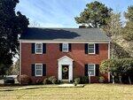 A two-story red brick house with black shutters, white trim, and a centered white portico over the front door. Neatly trimmed shrubs and a small lawn line the walkway leading to the entrance.