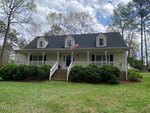 A two-story house with light-colored siding, a dark shingled roof, three dormer windows, a front porch with white railings, steps leading to the entrance, an American flag, and trimmed bushes in the front yard.