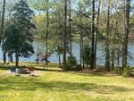 A grassy yard with several trees overlooks a calm river. In the foreground, four colorful chairs are arranged around a small fire pit. The setting is peaceful and natural with sunlight filtering through the trees.