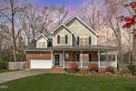 Two-story suburban house with beige siding and red brick exterior, white trim, double garage, covered front porch, and a manicured lawn, set against a backdrop of tall trees and a colorful sunset sky.