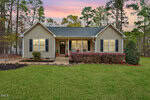 A single-story house with tan siding, black shutters, and a front porch, surrounded by a neatly trimmed lawn and bushes. Tall trees and a pink and purple sunset sky are in the background.