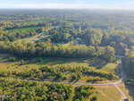 Aerial view of a rural landscape with a mix of open grassy fields, clusters of trees, and a winding dirt road. Residential houses and tree-covered areas are visible in the background under a clear sky.
