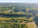 Aerial view of a rural landscape featuring a lightly wooded area, open grassy fields, a winding dirt road, and a small residential neighborhood surrounded by dense trees. The sky is clear with some distant haze.