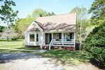 A small, light-colored house with a steep roof, front porch, and bay window sits on a grassy lot with trees in the background and a gravel driveway in the foreground. A flagpole stands near the porch.