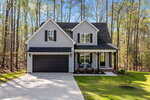 A two-story gray house with black shutters and a black garage door, featuring a covered front porch with two chairs, a small lawn, and surrounded by tall trees.