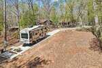 A large RV is parked on a paved campsite in a wooded area with a picnic table nearby. Trees surround the site, and several cabins or houses are visible in the background. The scene suggests a quiet, natural campground.